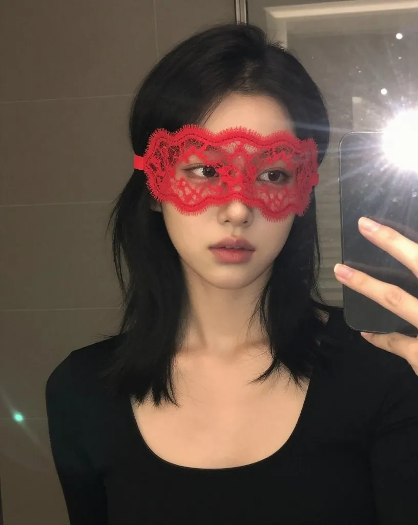붉은 레이스 가면을 쓰고 긴소매 검은 옷 차림으로 거울 앞에서 휴대폰을 든 여성